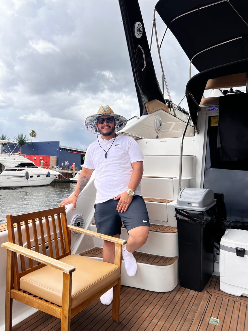Cristian en la cubierta de un yate con sombrero de paja y camiseta blanca, con la marina detrás